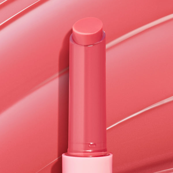Guava Glaze shade of the ColourPop So Juicy Plumping Gloss Balm with Peptides, a twist‑up coral‑pink balm standing upright with the bullet extended, showing a glossy finish against a matching guava‑pink swirled backdrop.
