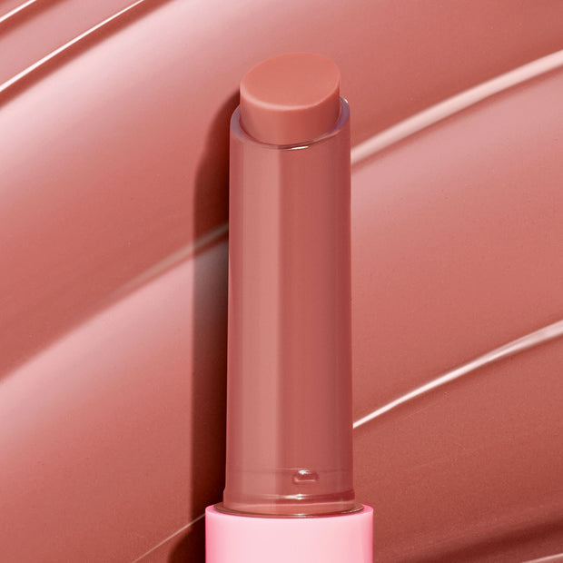 ColourPop So Juicy Plumping Gloss Balm with Peptides in Vacay Mode stands uncapped with the bullet extended, a warm rosy nude shade, in a pink tube against a matching glossy swatch background.