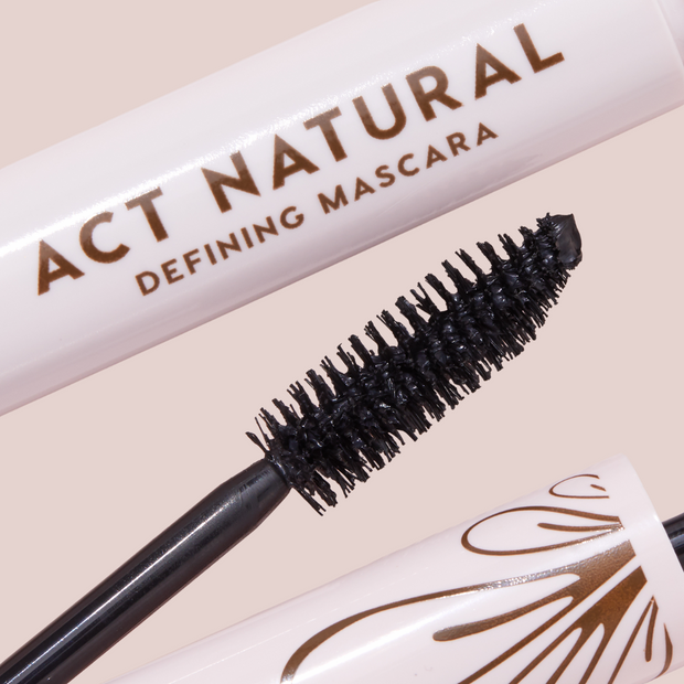 Mascara wand coated in deep black formula defining dense bristles, shown beside a blush-pink tube reading ACT NATURAL; ColourPop Act Natural Mascara, Black shade, with bronze detailing on the cap, close-up product shot on a neutral background.