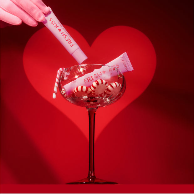 ColourPop Fresh Kiss lip gloss tubes being placed into a coupe glass filled with red-and-white peppermint candies and a mini candy cane, set against a bold red backdrop with a glowing heart for a festive holiday beauty scene.