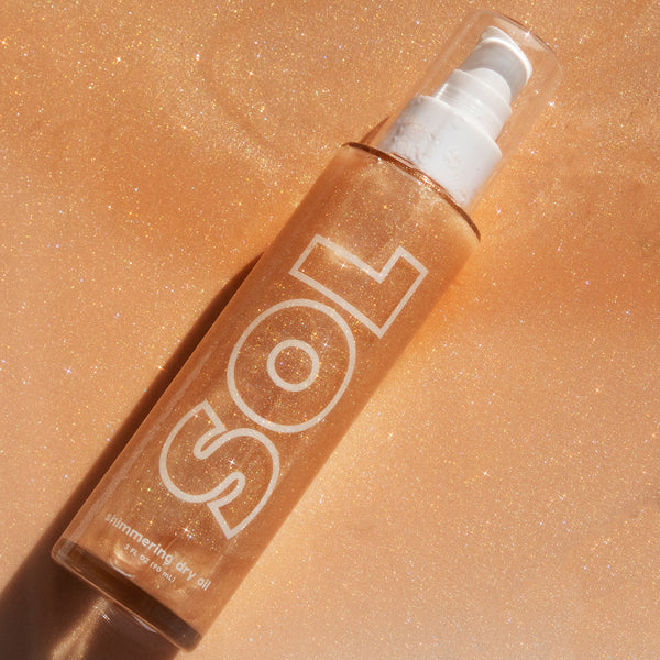 ColourPop SOL Shimmering Dry Oil in Warm Gold lying diagonally, a clear pump bottle with bold SOL lettering sparkling with fine gold shimmer, 3 fl oz/90 mL, set on a glittering golden backdrop that emphasizes its radiant body glow.