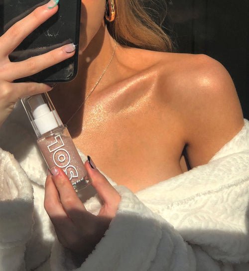 ColourPop SOL Shimmering Dry Oil in Soft Pink - clear glass bottle with white pump and pink shimmer - held near a sunlit collarbone glowing to show the luminous finish; close-up mirror shot in a cozy white robe.