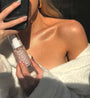ColourPop SOL Shimmering Dry Oil in Soft Pink - clear glass bottle with white pump and pink shimmer - held near a sunlit collarbone glowing to show the luminous finish; close-up mirror shot in a cozy white robe.