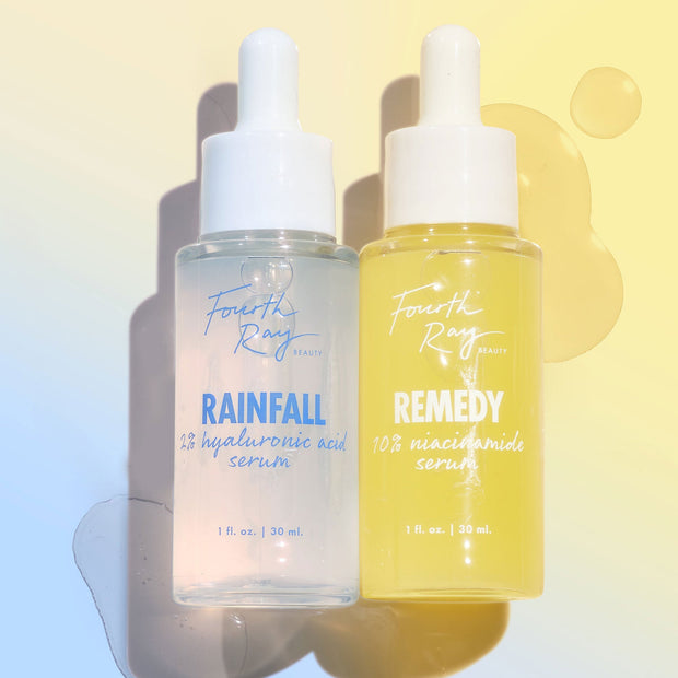 Remedy + Rainfall duo by Fourth Ray Beauty, two dropper bottles - Rainfall 2% hyaluronic acid serum and Remedy 10% niacinamide serum - standing side by side with glossy droplets on a pastel yellow-to-blue backdrop; ColourPop Face Serum set, 1 fl oz each.