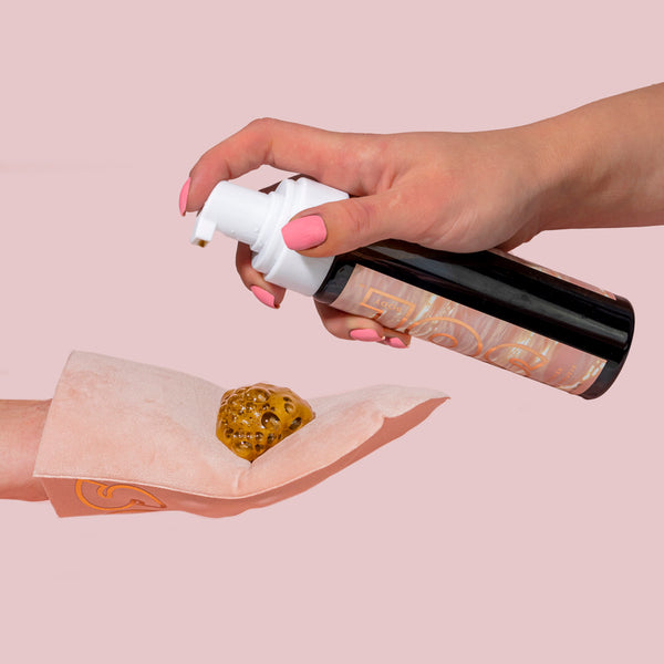 Tanning Mitt catching a pump of golden tanning foam from a black pump bottle, illustrating application for a streak-free sunless tan on a blush pink background, part of ColourPop Makeup Tools & Accessories.