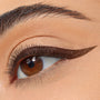 Winged eyeliner defines the upper lash line in a sharp, deep-brown cat eye, created with ColourPop BFF Liquid Liner in Grande, on a close-up of a brown eye with soft nude shadow and a groomed brow.