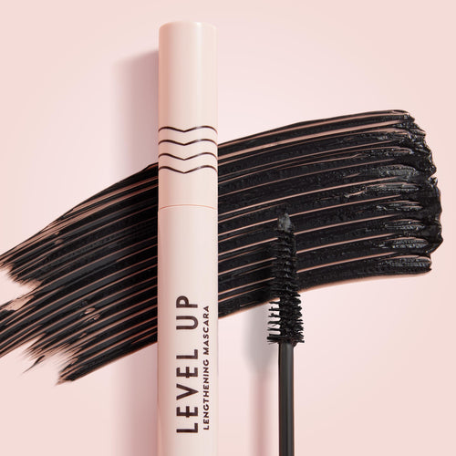 ColourPop Lengthening Mascara Level Up in Black stands upright with its wand coated in pigment, set before bold black swipes on a soft pink background, highlighting the slim brush and blush-pink tube for dramatic lash length.
