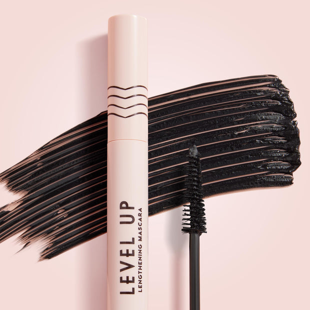 ColourPop Lengthening Mascara Level Up in Black stands upright with its wand coated in pigment, set before bold black swipes on a soft pink background, highlighting the slim brush and blush-pink tube for dramatic lash length.