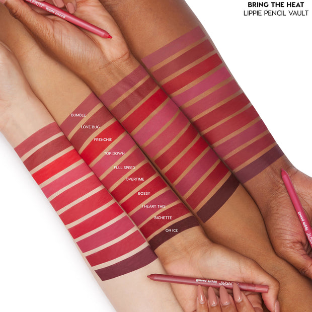 ColourPop Lippie Vault Set Heat Warning swatches stripe several arms, showcasing red, pink, and berry lippie pencil shades - Bumble, Love Bug, Frenchie, Bossy, I Heart This, Bichette, On Ice - while hands hold matching pencils on a white studio backdrop.