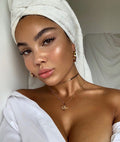 Warm Gold ColourPop SOL Glow Oil illuminates a model’s cheeks, collarbones, and shoulders with a dewy, sun-kissed sheen; towel-wrapped hair, white off-shoulder shirt, and gold jewelry in soft natural light emphasize the glow.
