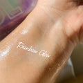 Rainbow Glow ColourPop SOL Shimmering Dry Oil swatched on inner wrist, catching light with iridescent rainbow micro-shimmer and a dewy sheen, shown close-up with soft-focus pastel packaging in the background.