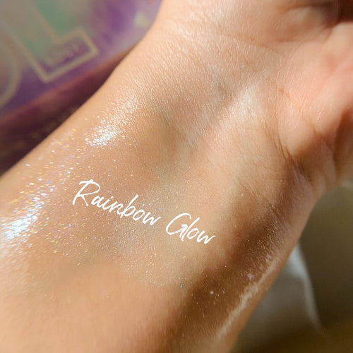 Rainbow Glow ColourPop SOL Shimmering Dry Oil swatched on inner wrist, catching light with iridescent rainbow micro-shimmer and a dewy sheen, shown close-up with soft-focus pastel packaging in the background.
