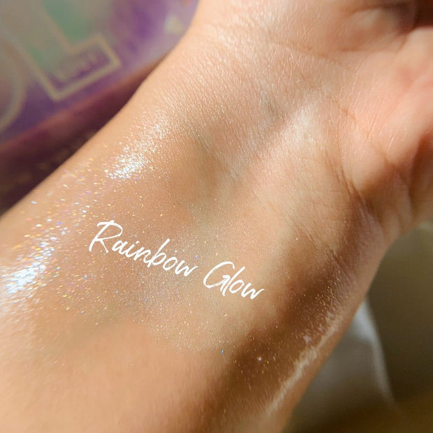 Rainbow Glow ColourPop SOL Shimmering Dry Oil swatched on inner wrist, catching light with iridescent rainbow micro-shimmer and a dewy sheen, shown close-up with soft-focus pastel packaging in the background.