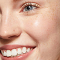 Lashes coated with ColourPop Act Natural Mascara in Brown, gently lengthening and separating a blue eye; close-up of a smiling face with freckles and dewy skin, showcasing a soft, natural brown mascara finish.