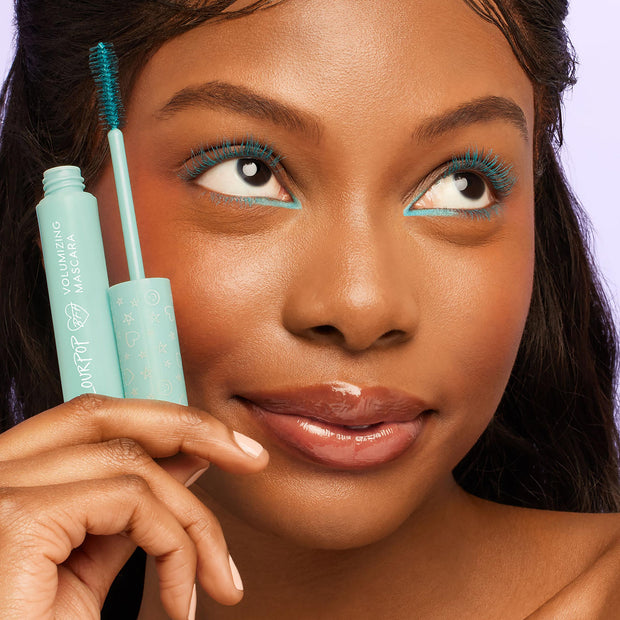 ColourPop BFF Mascara Swell Season paints lashes a bold teal as a model holds the mint volumizing mascara tube and brush near her eye, showing the star-printed cap, matching teal liner, and a soft lavender background.