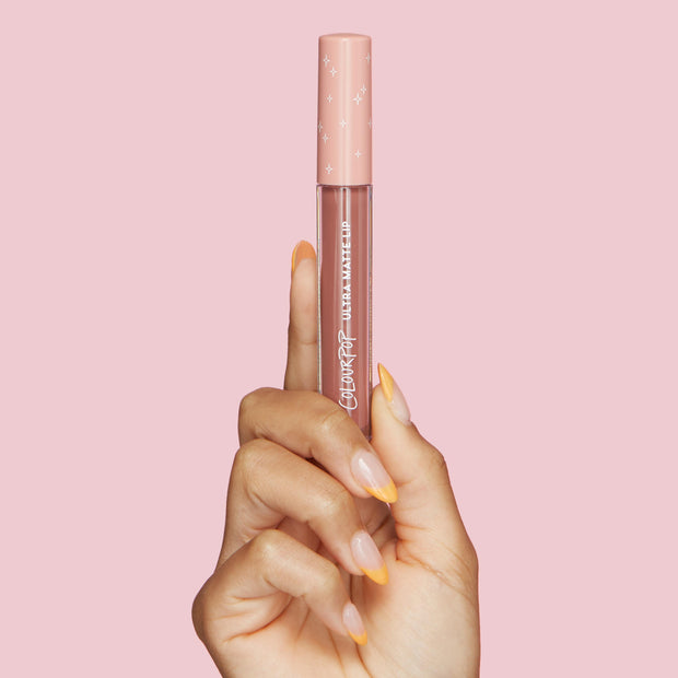 ColourPop Ultra Matte Lip in Beeper, a mauve‑nude liquid lipstick, held upright by a manicured hand with peach‑tipped nails; clear tube shows the shade and a peach cap with white stars against a soft pink background.