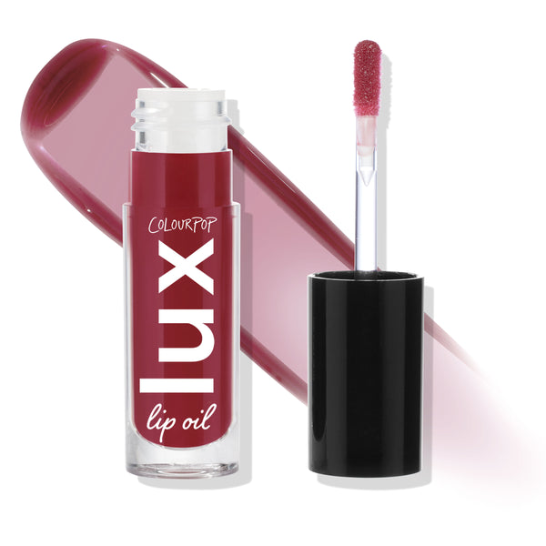 ColourPop Lux Lip Oil in Bells stands uncapped with a doe-foot applicator and black cap, showcasing a glossy berry-mauve tint; the clear tube displays Lux lip oil branding beside a matching swatch on a white background.
