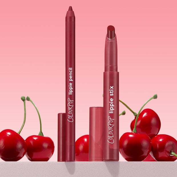 Bichette ColourPop Lippie Pencil and matching Lippie Stix stand upright with caps off, revealing rich cherry-red tips, surrounded by glossy cherries on a pink gradient backdrop, highlighting a creamy, precise liner duo for bold red lips.