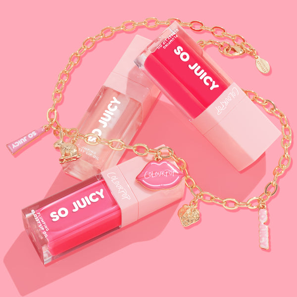 ColourPop So Juicy Lip Oil and Necklace Set, Charmed and Juicy, showcasing three plumping lip oils in clear and hot pink tubes, draped with a gold charm necklace featuring lip, heart, bunny, and logo charms on a pink flat lay.