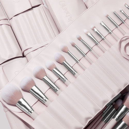 Stone Cold, a ColourPop Makeup Brush Kit, fanned in a blush-pink roll as face and eye brushes slot into stitched pockets, soft white-to-pink bristles and silver ferrules gleaming, with additional matching brush rolls layered behind.