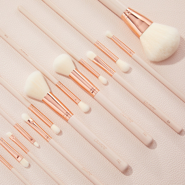 ColourPop Makeup Brush Kit lined up diagonally, the Ultimate Brush Roll features fluffy face and eye brushes with white bristles, rose-gold ferrules, and pale pink handles resting on a blush textured backdrop.