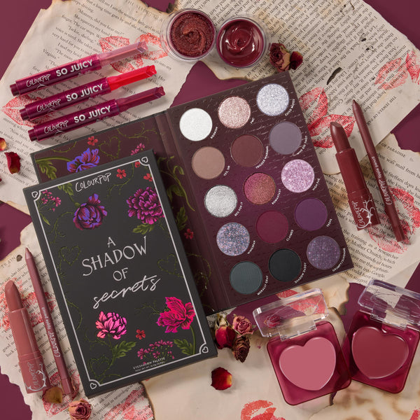 ColourPop Full Collection Set A Shadow of Secrets Collection arranged in a top-down flat lay, featuring a floral box, berry-toned eyeshadow palette, heart-shaped blushes, So Juicy glosses, lipsticks, liners, and cream pots on torn pages with rose petals.