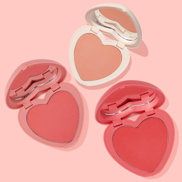 ColourPop Heart Powder Blush Set Cupid Called displays three heart-shaped powder blush compacts opened to reveal peach nude, coral pink, and bright pink shades with mirrors, arranged diagonally on a soft pink background in white and pink cases.