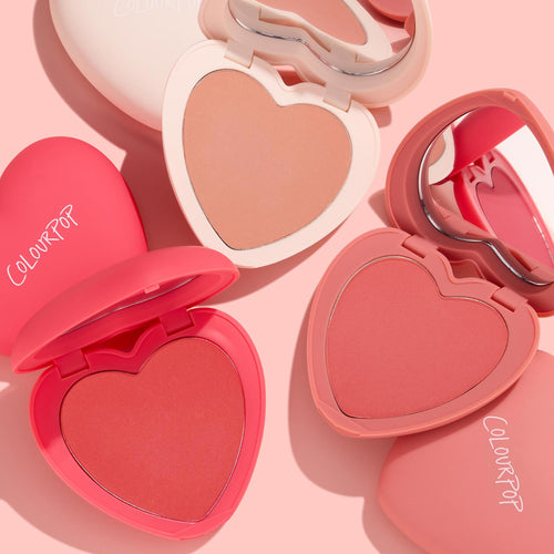 Cupid Called by ColourPop Heart Powder Blush Set opens to reveal three heart-shaped pans - peach, coral, and rosy pink - with matching mirrored compacts, arranged in a flat lay on a soft pink backdrop.