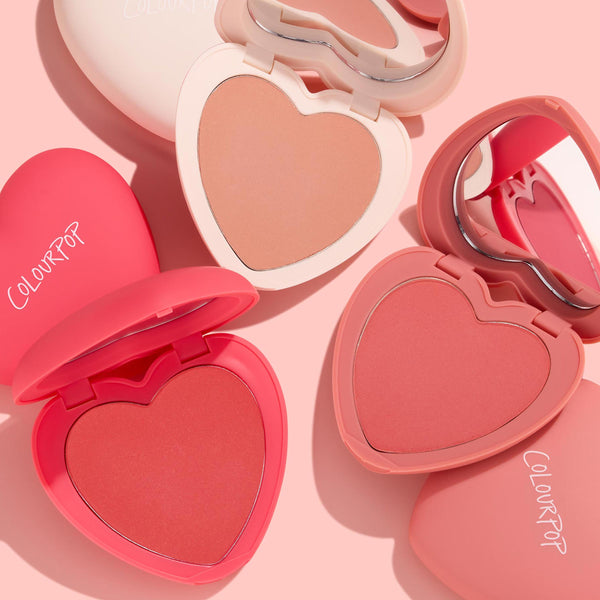 Cupid Called by ColourPop Heart Powder Blush Set opens to reveal three heart-shaped pans - peach, coral, and rosy pink - with matching mirrored compacts, arranged in a flat lay on a soft pink backdrop.