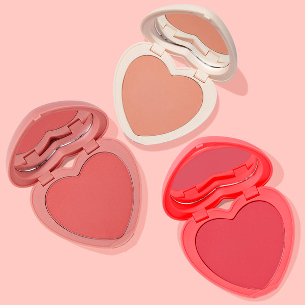 Current Crush by ColourPop Heart Powder Blush Set showcasing three heart-shaped powder blush compacts opened to reveal mirrors and matte pans, in peach, coral, and rosy pink shades with white, soft pink, and neon coral cases on a pastel pink backdrop.