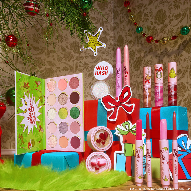 ColourPop x The Grinch Full Collection Set showcasing an open 15‑shade eyeshadow palette, lip crayons, lip glosses, and Super Shock shadows with heart cutouts, arranged on wrapped gifts in a festive holiday scene; playful pink‑green packaging, ColourPop collab.