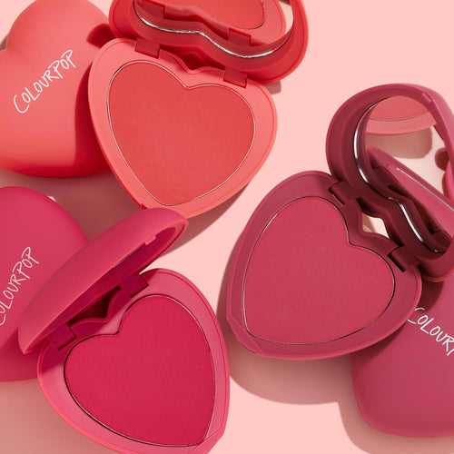 Heart to Heart blush compacts displaying three heart-shaped powder pans in coral, rose, and berry, opened with mirrors on a soft pink surface, ColourPop branding on pink cases; ColourPop Heart Powder Blush Set.