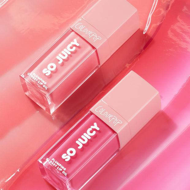 Two ColourPop So Juicy Lip Oil bottles from the It's Giving Juicy bundle lie diagonally, showing rosy-pink shades in clear square tubes with blush caps and SO JUICY plumping glossy lip oil text on a glossy pink backdrop; ColourPop So Juicy Lip Oil Set.