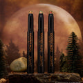 Three Love Triangle ColourPop Shadow Stix stand upright, their shimmery duochrome cream tips glowing green-gold and pink, displayed on a rocky surface with pine trees and a giant full moon backdrop for a moody, twilight woodland vibe.