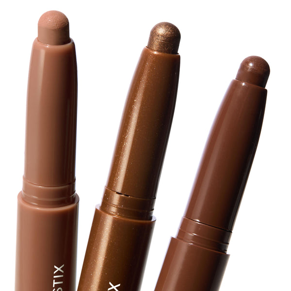 Three ColourPop Shadow Stix Essential Neutrals stand uncapped, revealing twist-up cream eyeshadow tips in nude beige, metallic bronze shimmer, and deep chocolate brown, arranged close-up on a clean white background.
