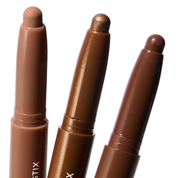 Three ColourPop Shadow Stix Essential Neutrals stand uncapped, revealing twist-up cream eyeshadow tips in nude beige, metallic bronze shimmer, and deep chocolate brown, arranged close-up on a clean white background.