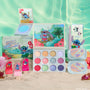 Disney Lilo and Stitch Collection by ColourPop Full Collection Set displayed on sandy beach - 12-pan Beach Dreamin’ eyeshadow palette, lip gloss trio, two cream gel liners, marbled highlighter compact, and jelly highlighter in tropical Stitch packaging.