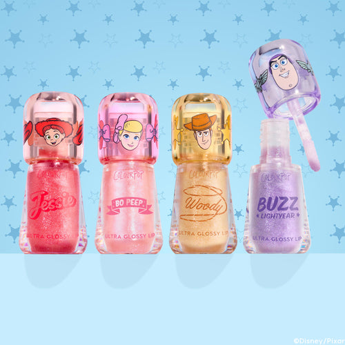 Squad Dreams ColourPop Ultra Glossy Lip bundle lined up: Jessie coral pink shimmer, Bo Peep soft pink shimmer, Woody warm gold glitter, Buzz Lightyear lavender sparkle with wand lifted; character caps; light blue Toy Story sheriff-star backdrop.