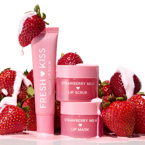 Strawberry Feels ColourPop Lip Care Kit displays a pink Fresh Kiss glossy lip balm beside stacked Strawberry Milk lip scrub and lip mask, dripping with strawberry milk and surrounded by ripe strawberries on a clean white backdrop.
