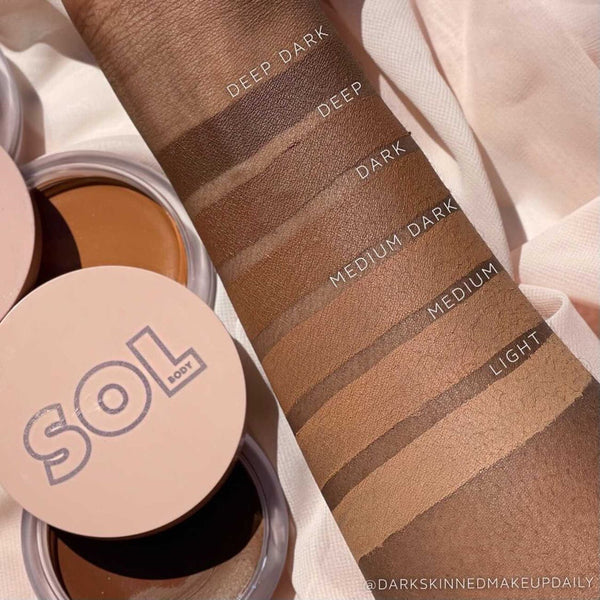 ColourPop SOL Face & Body Bronzing Balm, Fair shade compact, sitting open beside an arm swatched and labeled Light, Medium, Medium Dark, Dark, Deep, and Deep Dark on soft fabric, showing the cream bronzer’s shade range and finish.