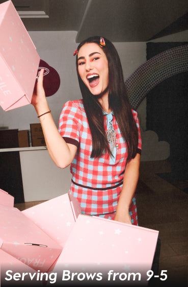 Smiling woman in a red and blue checkered dress excitedly lifting a pink brow product shipping box in a playful studio packed with more branded pink packages, with the slogan “Serving Brows from 9-5” along the bottom