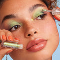 See U Later ColourPop Chrome Liquid Shadow is applied to the eyelid with a doe-foot applicator, revealing a lime-to-gold shift; close-up shows the ColourPop-labeled mini bottle near glossy lips, coral nails, freckles, and a sky-blue background.