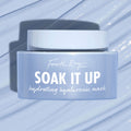 Fourth Ray Beauty Soak It Up Hydrating Mask jar shows “hydrating hyaluronic mask” as it rests on a glossy, water-swept pale blue surface; frosted periwinkle square container with white lid, a ColourPop Face Mask for dewy hydration.