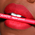 ColourPop Lippie Pencil in Overtime held between vividly lined, matte red lips, showcasing the creamy lip liner’s precise definition in a close-up beauty shot with the hot-pink barrel and white logo text visible.