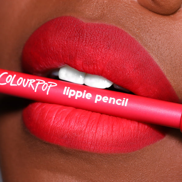 ColourPop Lippie Pencil in Overtime held between vividly lined, matte red lips, showcasing the creamy lip liner’s precise definition in a close-up beauty shot with the hot-pink barrel and white logo text visible.