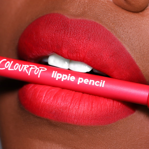 ColourPop Lippie Pencil in Overtime held between vividly lined, matte red lips, showcasing the creamy lip liner’s precise definition in a close-up beauty shot with the hot-pink barrel and white logo text visible.