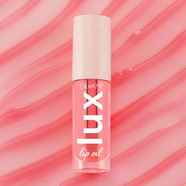 ColourPop Lux Lip Oil in Cherry Bite stands upright, displaying a clear pink-tinted tube with bold white LUX logo and a cream cap patterned with stars, set against glossy pink swirls that echo the coral lip oil tint.