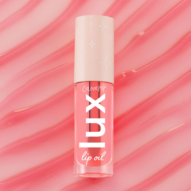 ColourPop Lux Lip Oil in Cherry Bite stands upright, displaying a clear pink-tinted tube with bold white LUX logo and a cream cap patterned with stars, set against glossy pink swirls that echo the coral lip oil tint.
