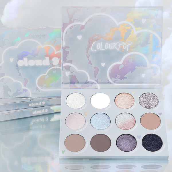 Cloud 9 ColourPop Shadow Palette opens to reveal 12 cool-toned neutrals and iridescent shimmers - from icy silver and matte white to taupe, lilac, and deep navy - set in holographic cloud packaging with stacked palettes and pastel clouds behind.