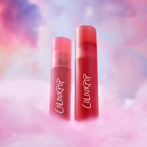 ColourPop Sunset Smooch Lip Ombre Duo standing upright as a red and pink liquid lipstick value set, rising from fluffy pink clouds against a dreamy pastel sky background.